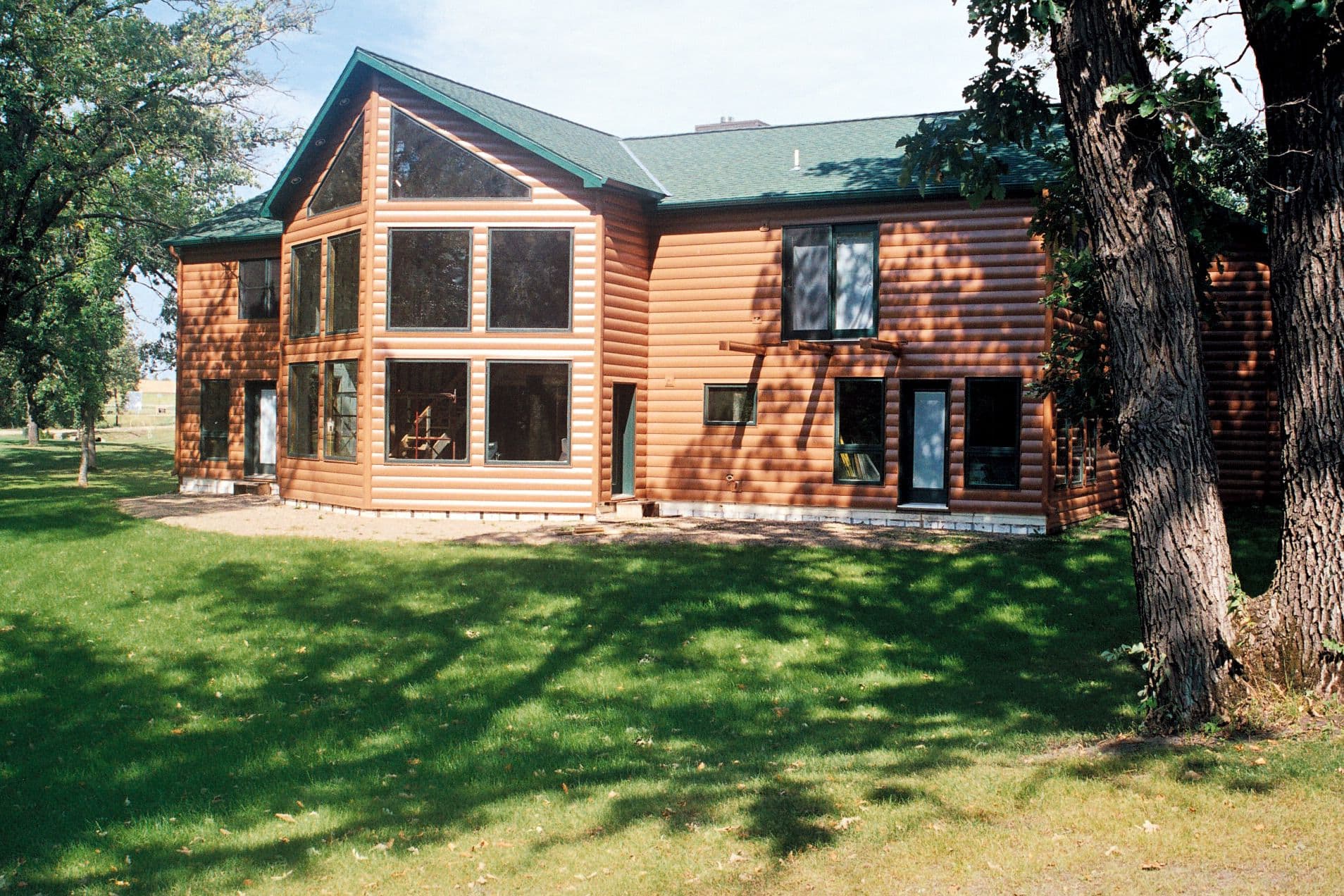Two-story log cabin with green metal roof and multiple windows, situated on a grassy lawn with large trees nearby