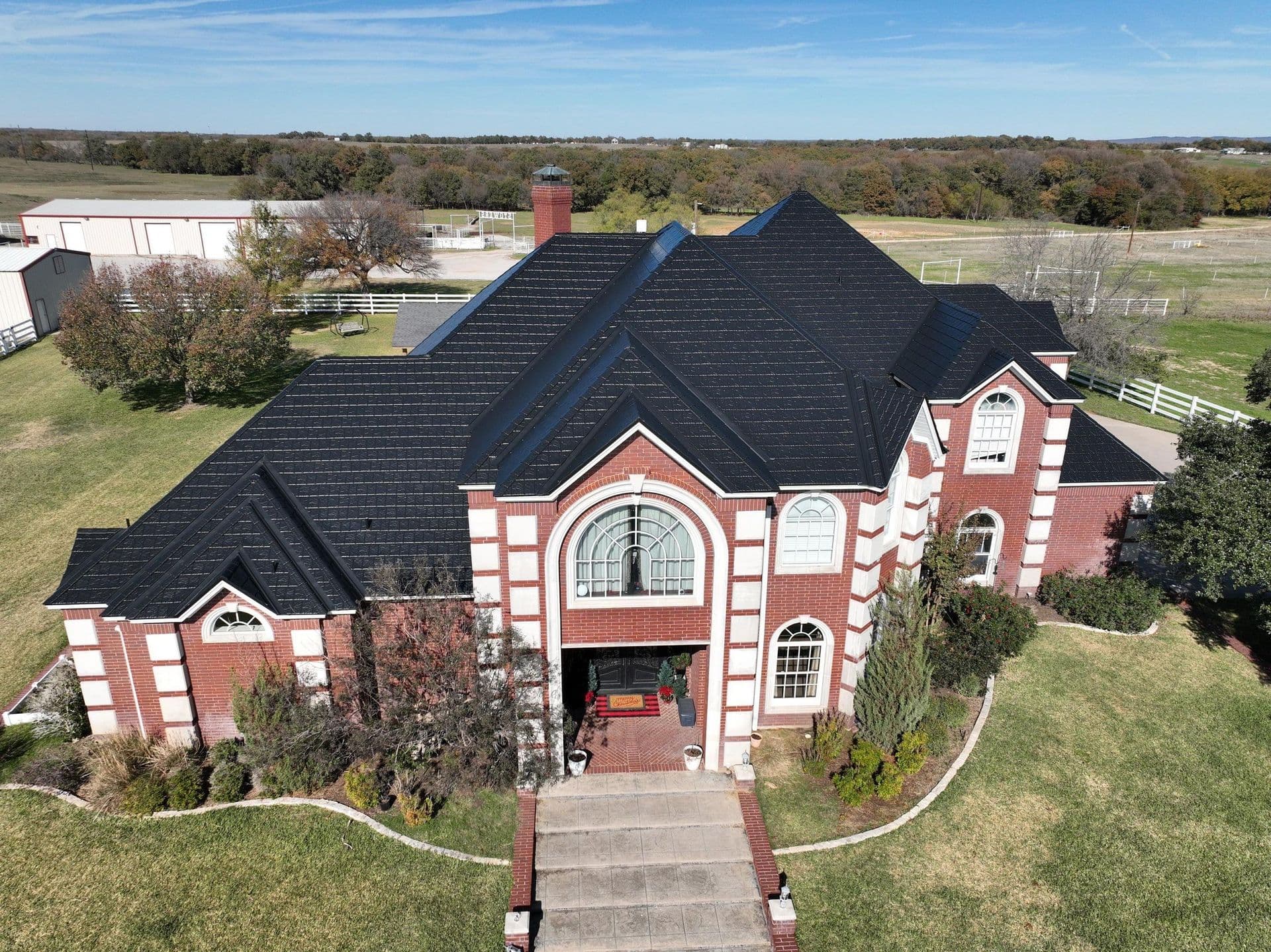 Aerial view of a two-story red brick house with black roof, circular window, two-car garage, and landscaped grounds on rural property with farm buildings and wooded areas in background