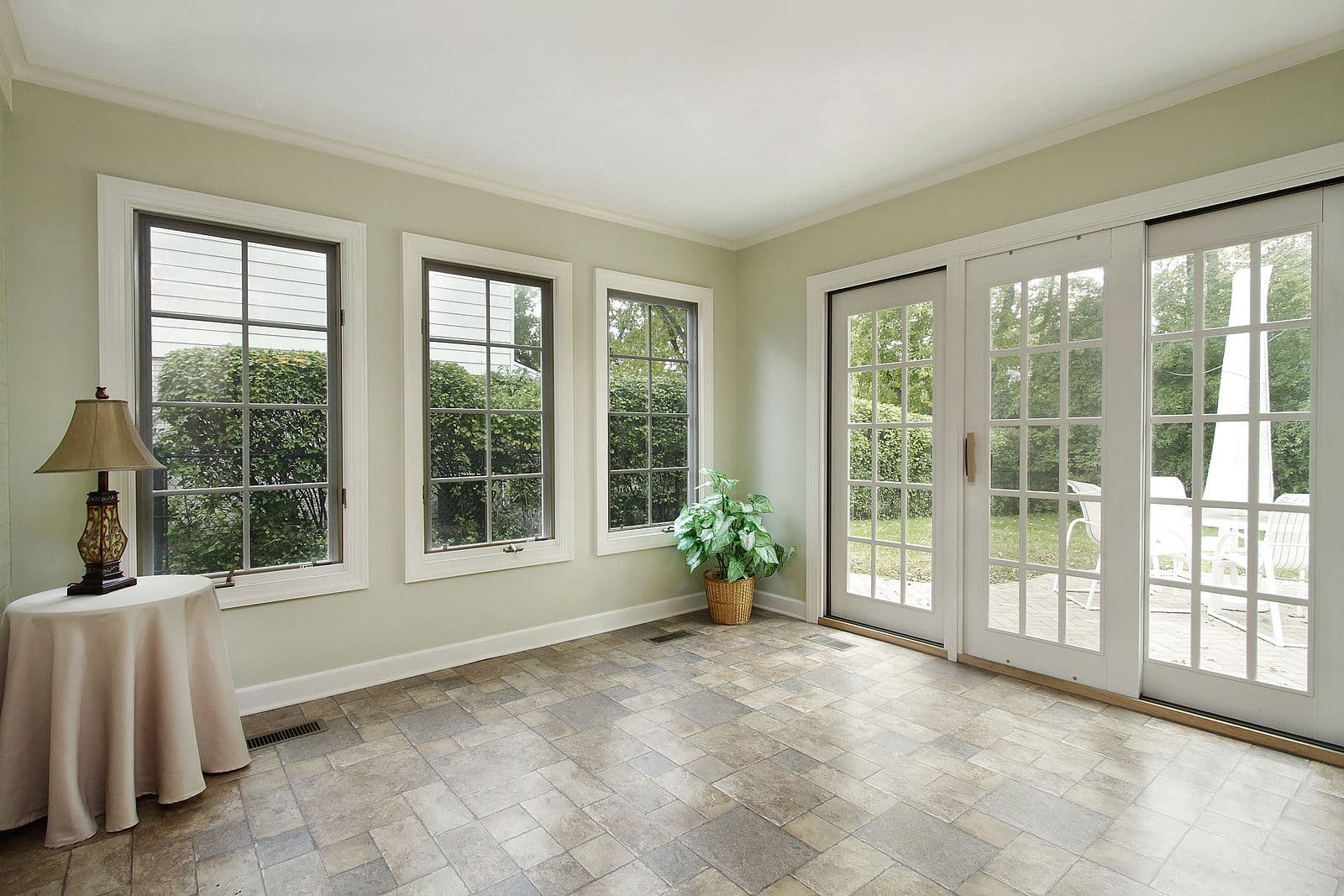 Sunlit conservatory with pale green walls, multiple windows and glass doors overlooking garden, stone tile flooring, side table with lamp and potted plant
