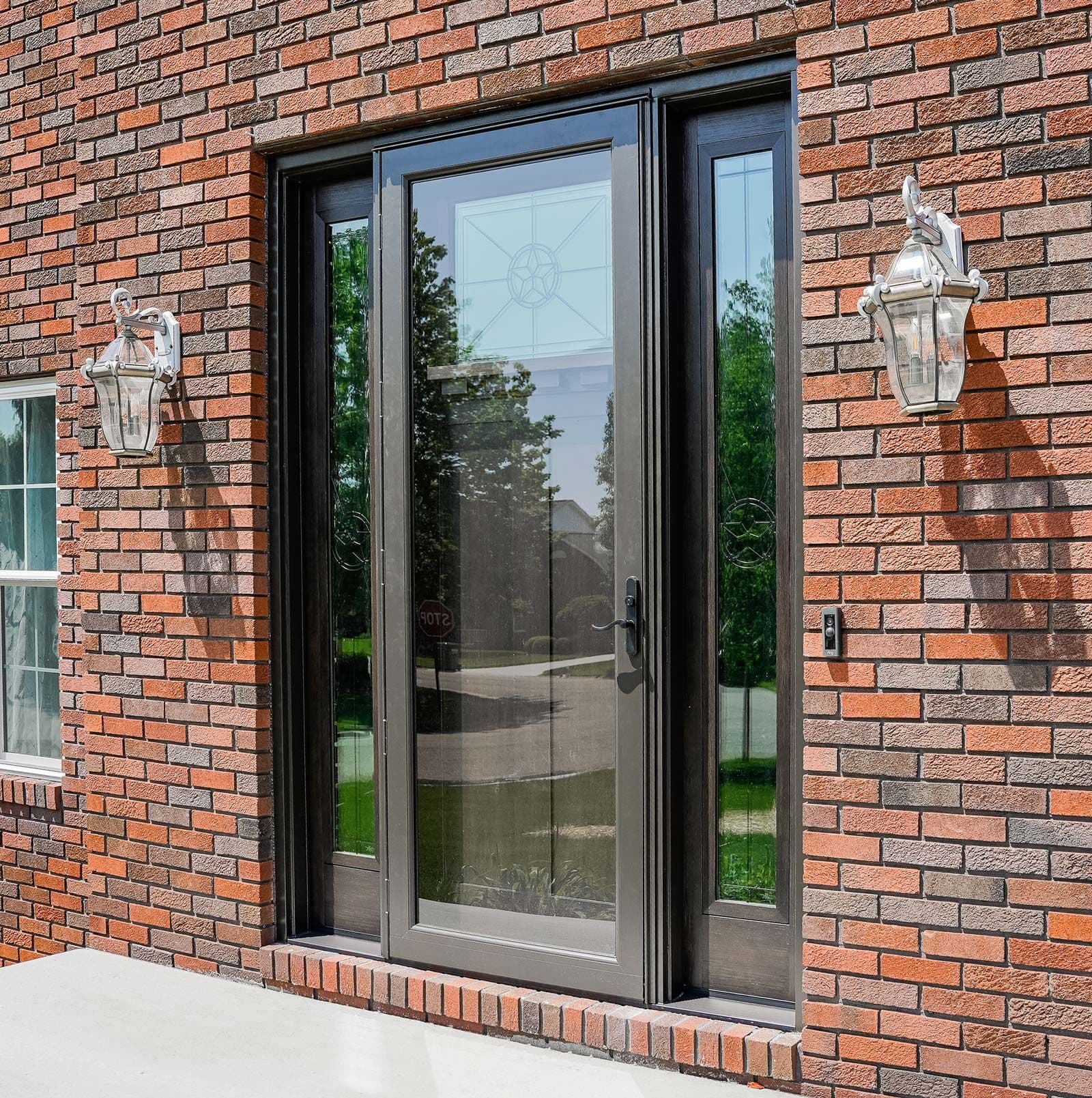 Black-framed glass patio door with sidelights on red brick exterior wall, flanked by white lantern fixtures