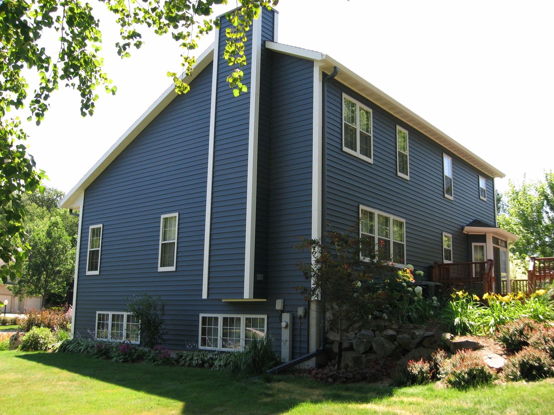 Two-story blue house with white-trimmed windows, elevated foundation, wooden deck, and landscaped yard with green lawn and flowering shrubs