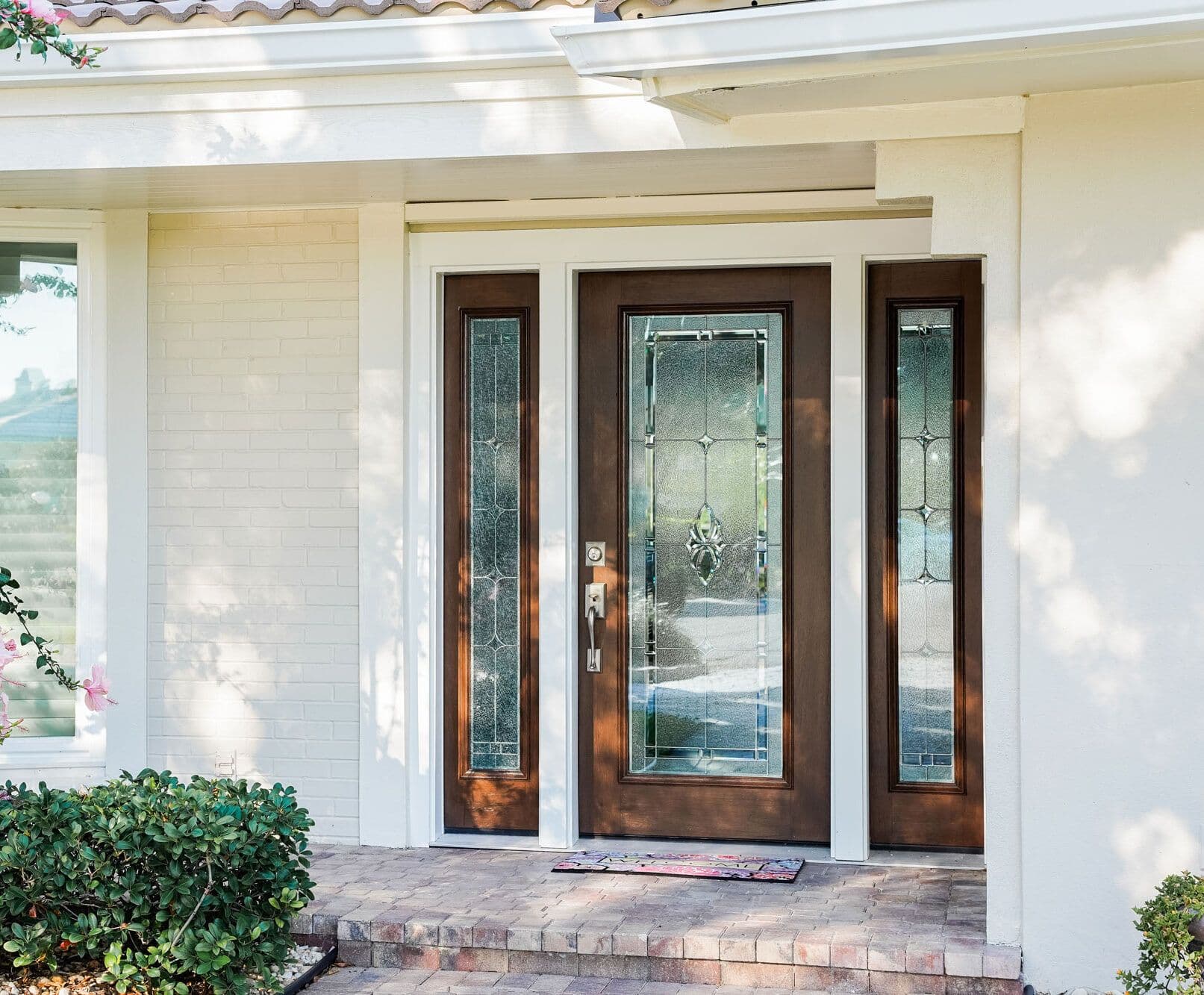 Front entrance of home with dark wood center door and matching sidelights featuring decorative glass panels, flanked by white brick walls and landscaping