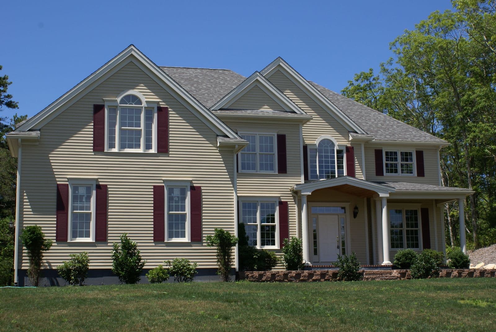 Two-story colonial house with tan siding, burgundy shutters, multiple peaked roofs, columned front porch, and brick foundation, surrounded by mature trees and green lawn