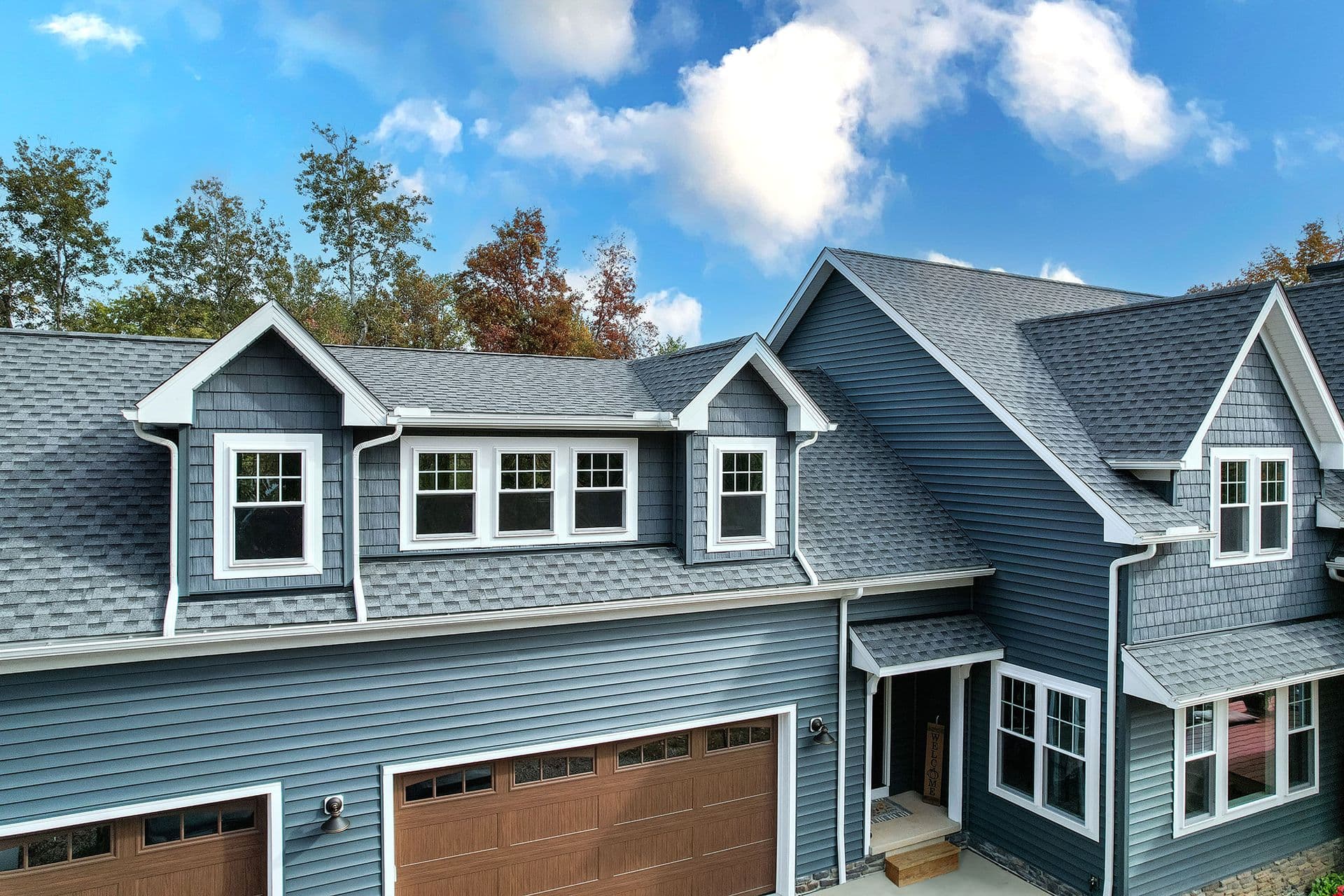 Modern two-story residential home with blue-gray siding, white trim, multiple gabled rooflines, dormer windows, and attached garage doors with decorative panels, surrounded by autumn trees under a partly cloudy sky